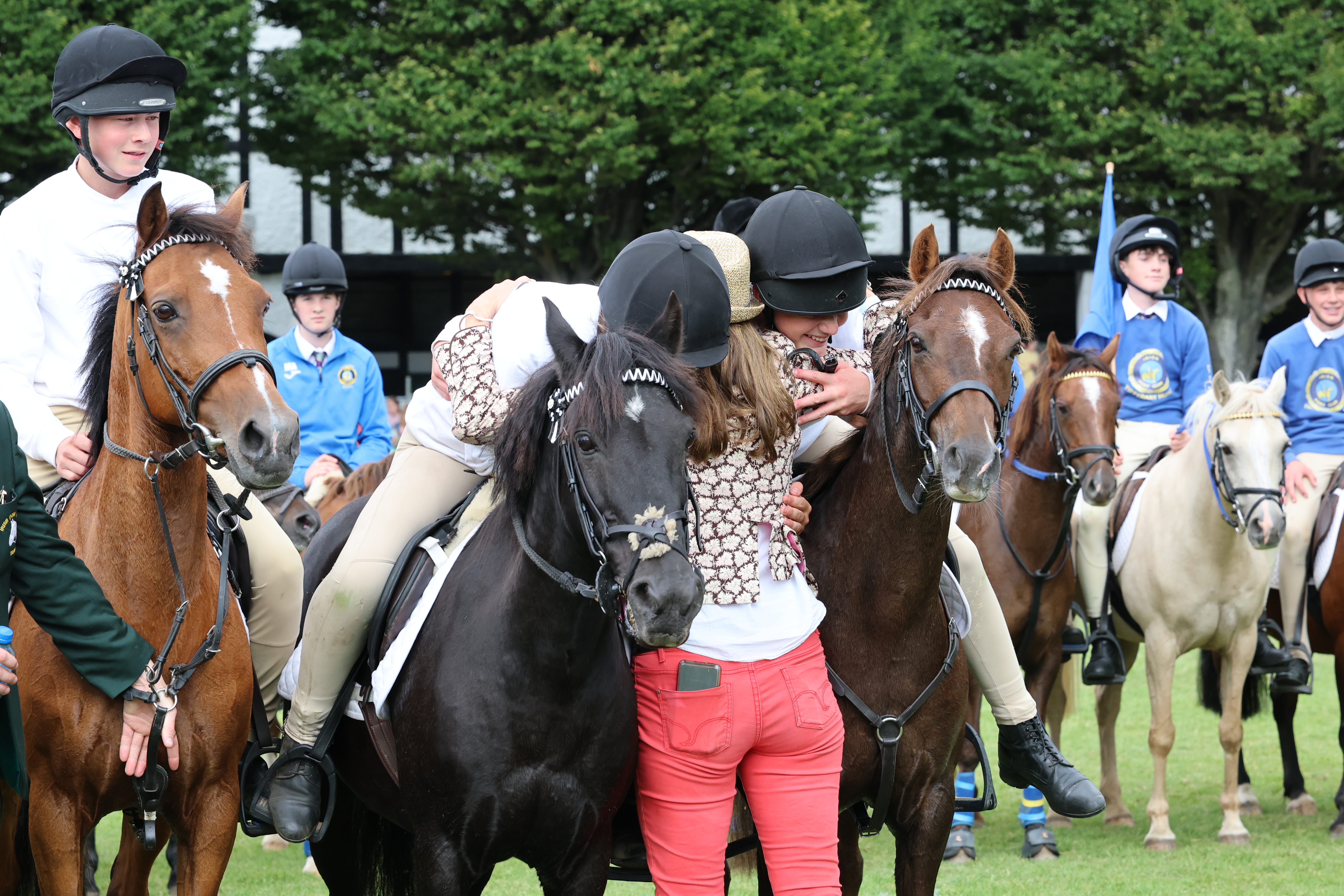 Une femme enlace deux jeunes cavaliers assis sur un poney brun et un poney noir.